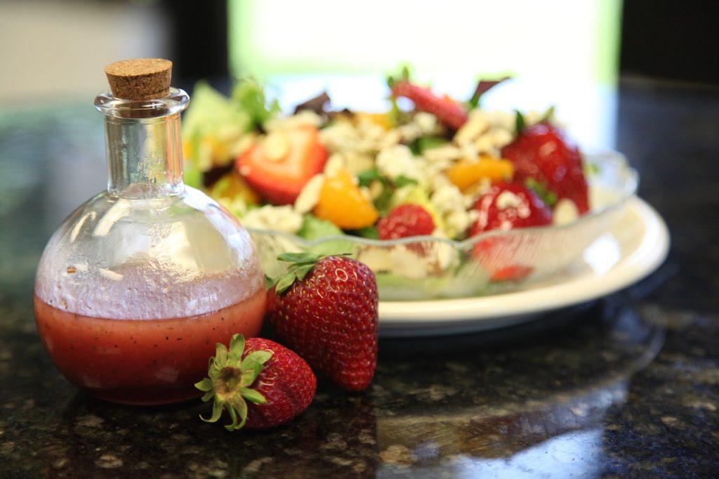 A corked glass bottle with salad dressing with two strawberries resting against it in the foreground and a glass bowl of salad in the background.