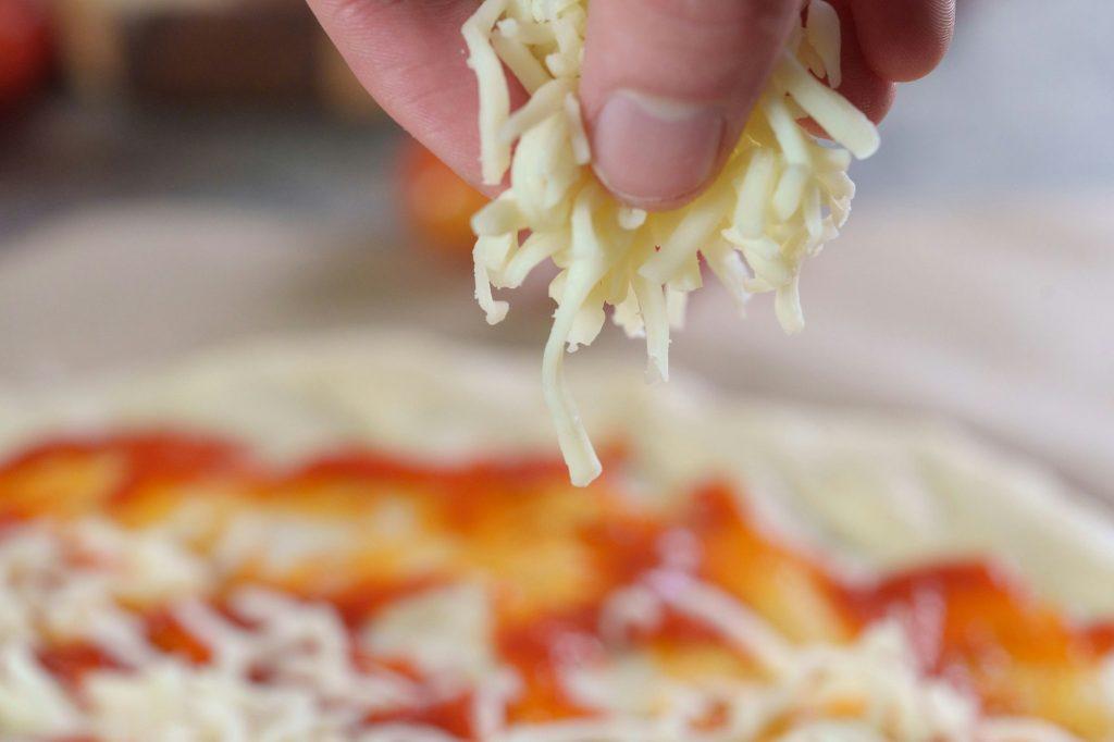 Person's hand adding grated cheese to pizza, process of making pizza